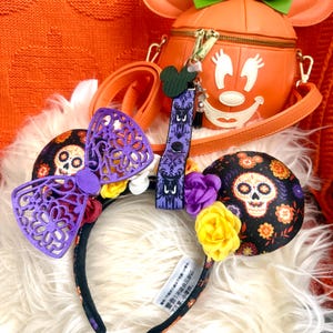 May include: An orange pumpkin-shaped handbag with a green bow, a Mickey Mouse keychain, and a headband with a purple bow and skull-themed ears. The headband has a black base with colorful floral accents.