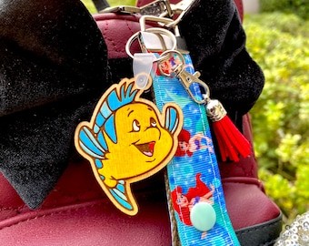 Puede incluir: Un amuleto de pez de madera amarillo y azul con una cara sonriente, unido a una cinta azul y blanca con imágenes de sirenas y burbujas. El amuleto está unido a un cordón rojo y blanco con una borla.