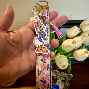 May include: A lanyard-style keychain featuring a Daisy Duck charm. The lanyard has a pink, blue, and yellow pattern with a gold-tone clasp and a pink tassel. The Daisy Duck charm is a cartoon character in a purple dress and pink bow.