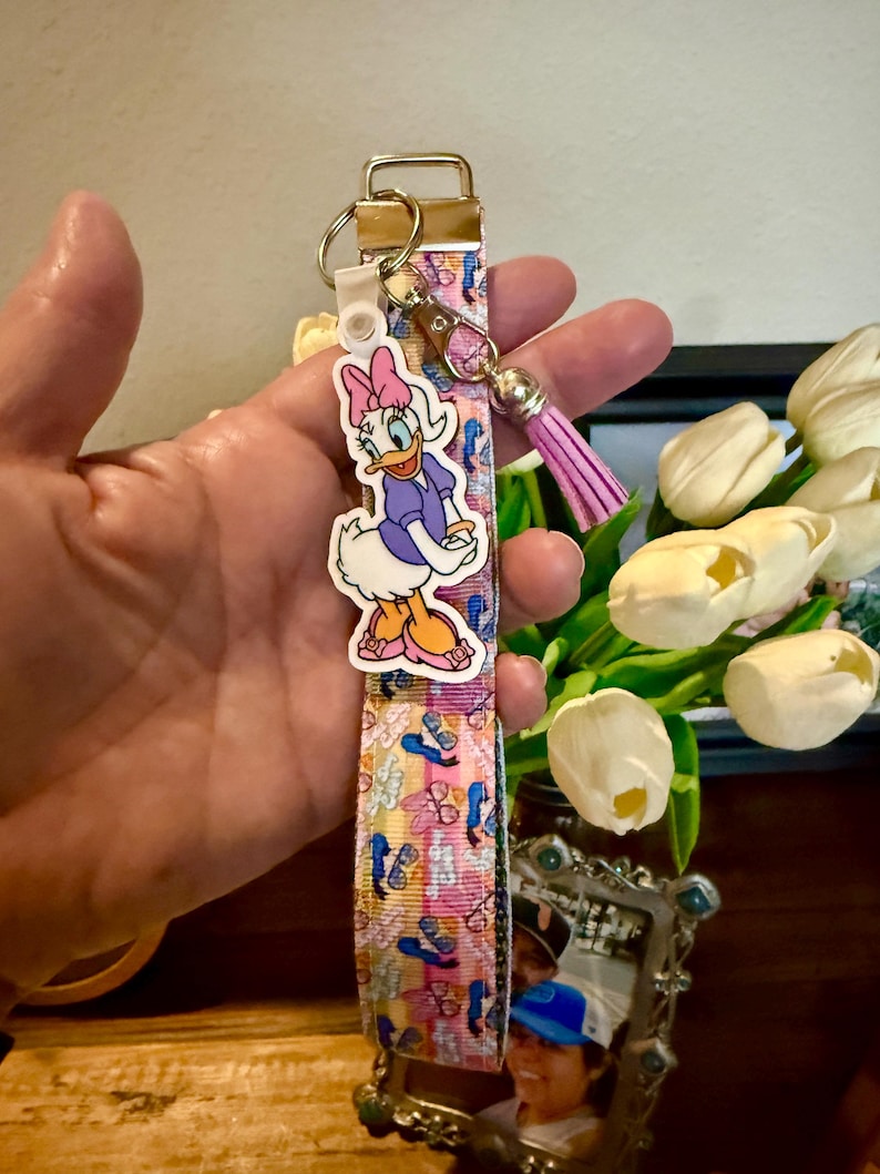 May include: A lanyard-style keychain featuring a Daisy Duck charm. The lanyard has a pink, blue, and yellow pattern with a gold-tone clasp and a pink tassel. The Daisy Duck charm is a cartoon character in a purple dress and pink bow.