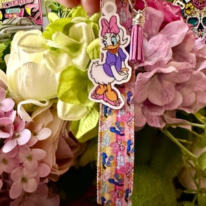 May include: A Daisy Duck keychain with a pink bow, purple dress, and orange shoes. The keychain strap features a colorful pattern of flip-flops and sunglasses. A pink tassel hangs from the keychain.