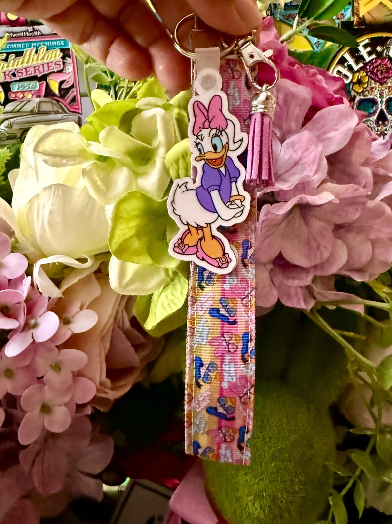 May include: A Daisy Duck keychain with a pink bow, purple dress, and orange shoes. The keychain strap features a colorful pattern of flip-flops and sunglasses. A pink tassel hangs from the keychain.