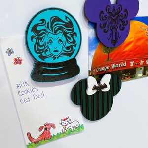 Op de afbeelding: Drie houten magneten met Disney-figuren. De eerste magneet is een turquoise blauwe sneeuwbol met een zwart silhouet van Ursula uit De Kleine Zeemeermin. De tweede magneet is een paarse Mickey Mouse-oorvorm met een zwart silhouet van het Haunted Mansion-logo. De derde magneet is een groene Mickey Mouse-oorvorm met zwarte strepen en een witte strik met een zwarte vleermuis.