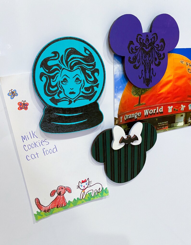 Op de afbeelding: Drie houten magneten met Disney-figuren. De eerste magneet is een turquoise blauwe sneeuwbol met een zwart silhouet van Ursula uit De Kleine Zeemeermin. De tweede magneet is een paarse Mickey Mouse-oorvorm met een zwart silhouet van het Haunted Mansion-logo. De derde magneet is een groene Mickey Mouse-oorvorm met zwarte strepen en een witte strik met een zwarte vleermuis.
