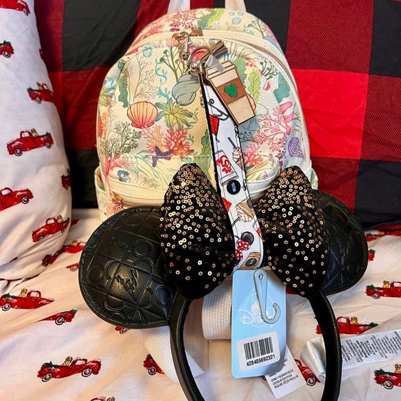 May include: A white and floral patterned mini backpack with a black and gold sequin Minnie Mouse headband. The headband has a black leather headband with a bow on top. The backpack has a zipper closure and a top handle. The backpack has a white and black strap with a Mickey Mouse coffee cup charm.
