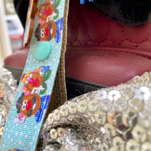 Pode incluir: Um close-up de uma fita azul clara com um padr&atilde;o repetido de personagens do Mr. Potato Head. Um bot&atilde;o verde claro est&aacute; preso &agrave; fita. A fita est&aacute; ao lado de um sapato vermelho e um tecido de lantejoulas prateadas.