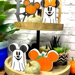 May include: Two-tiered wooden tray with a Halloween theme. The top tier features a black "LOVE" sign, a white ghost with a red bow, and a pumpkin with Mickey Mouse ears. The bottom tier features a white ghost with Mickey Mouse ears, a pumpkin with Mickey Mouse ears, and a small gold pumpkin.
