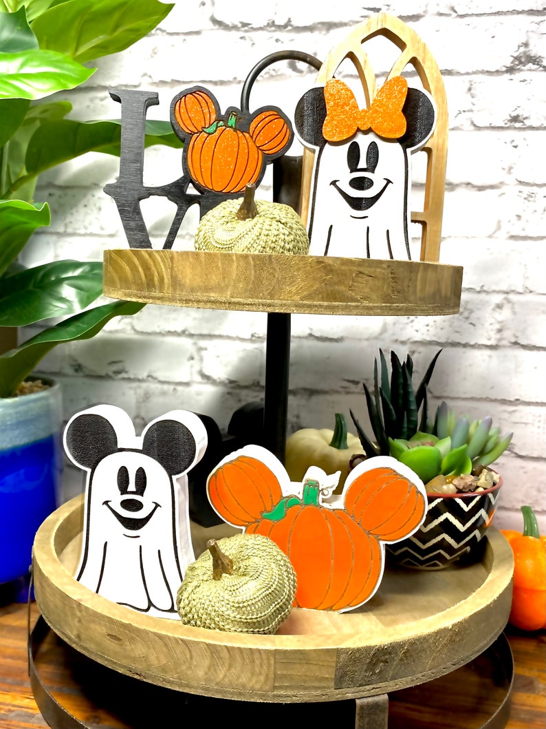 May include: Two-tiered wooden tray with a Halloween theme. The top tier features a black "LOVE" sign, a white ghost with a red bow, and a pumpkin with Mickey Mouse ears. The bottom tier features a white ghost with Mickey Mouse ears, a pumpkin with Mickey Mouse ears, and a small gold pumpkin.