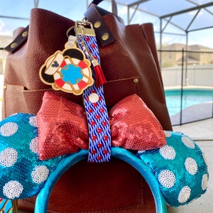 May include: A pair of blue and white sequined Minnie Mouse ears with red and white ribbon and a wooden life preserver charm with a red and white striped rope.