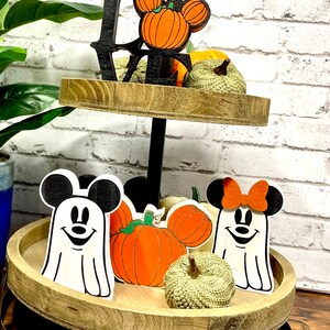 May include: A two-tiered wooden tray with a black metal stand. The top tier has a black wooden "LOVE" sign with a pumpkin-shaped Mickey Mouse head. The bottom tier has two ghost-shaped Mickey Mouse figures, a pumpkin, and a small green gourd.