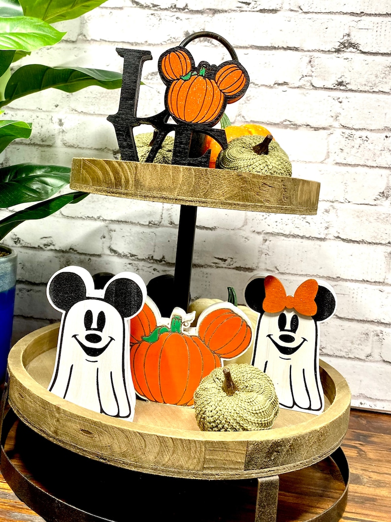 May include: A two-tiered wooden tray with a black metal stand. The top tier has a black wooden "LOVE" sign with a pumpkin-shaped Mickey Mouse head. The bottom tier has two ghost-shaped Mickey Mouse figures, a pumpkin, and a small green gourd.