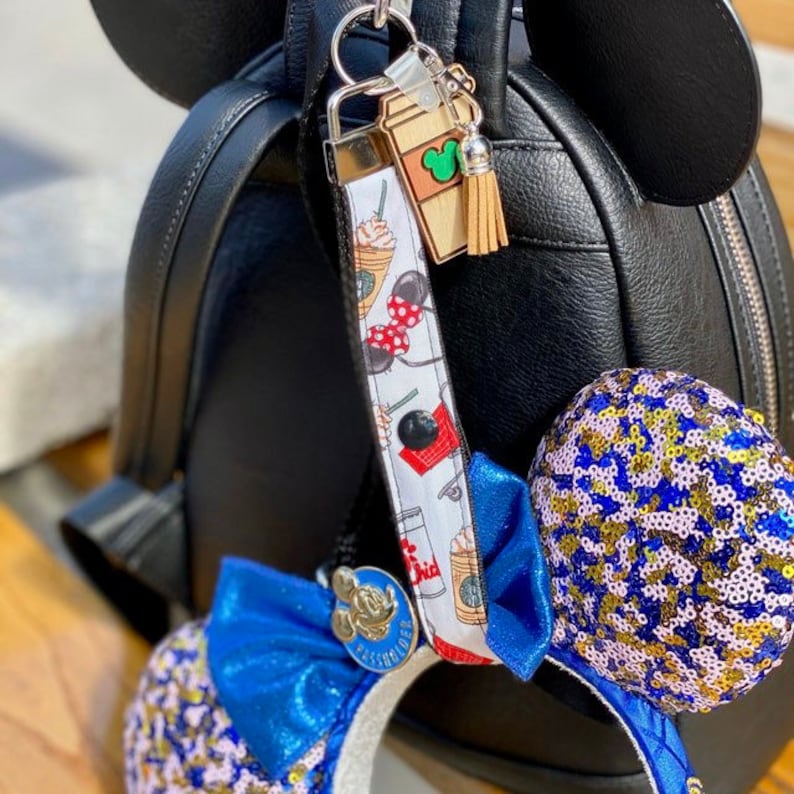Pode incluir: Uma mochila preta com orelhas do Mickey Mouse, um cord&atilde;o branco com um padr&atilde;o da Minnie Mouse e um pingente de x&iacute;cara de caf&eacute; de madeira com uma cabe&ccedil;a verde do Mickey. Uma tiara azul com lantejoulas e um la&ccedil;o azul tamb&eacute;m &eacute; vis&iacute;vel.