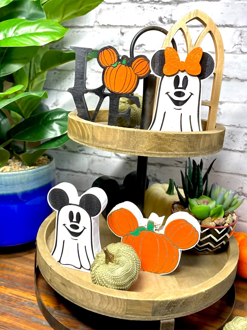 May include: A tiered wooden tray with Halloween decorations. The top tier has a black wooden "LOVE" sign with a pumpkin-shaped Mickey Mouse head. The bottom tier has a white ghost with Mickey Mouse ears, a pumpkin-shaped Mickey Mouse head, and a gold pumpkin.