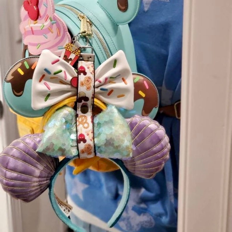 May include: A pair of purple and teal Minnie Mouse ears with a sparkly bow and a charm featuring a Mickey Mouse ice cream cone. The ears are attached to a headband.