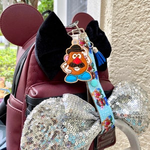 Pode incluir: Um chaveiro de madeira do Mr. Potato Head com um cord&atilde;o e borla azuis est&aacute; preso a uma mochila bord&ocirc; com la&ccedil;os pretos e uma tiara com la&ccedil;o de lantejoulas. O chaveiro apresenta um personagem de desenho animado com bigode e chap&eacute;u marrons.