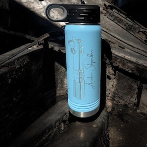 Puede incluir: Una botella de agua de acero inoxidable azul claro con tapa negra y asa de transporte. La botella tiene una imagen grabada con l&aacute;ser de un sable de luz y el texto "Anakin Skywalker".