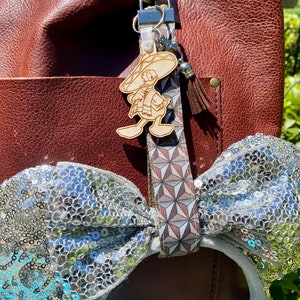 May include: A brown leather tote bag with a silver sequin bow headband and a wooden keychain featuring a cartoon character wearing a sombrero. The keychain is attached to a brown and white patterned ribbon.