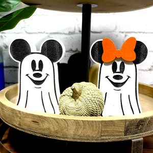 May include: Two wooden ghost figures of Mickey Mouse and Minnie Mouse, with black ears and eyes, and white faces. Mickey Mouse is wearing a white ghost costume and Minnie Mouse is wearing a white ghost costume with an orange bow. The figures are sitting on a wooden tiered tray with a small green pumpkin in front of them.