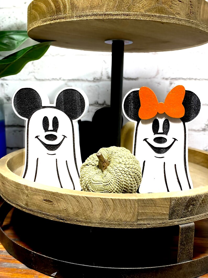 May include: Two wooden ghost figures of Mickey Mouse and Minnie Mouse, with black ears and eyes, and white faces. Mickey Mouse is wearing a white ghost costume and Minnie Mouse is wearing a white ghost costume with an orange bow. The figures are sitting on a wooden tiered tray with a small green pumpkin in front of them.