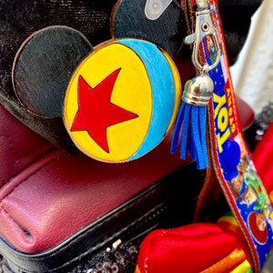 May include: A wooden keychain with a blue, yellow, and red design of a ball with a star. The keychain is attached to a brown lanyard with the words "Toy Story" printed on it. The lanyard has a blue tassel.
