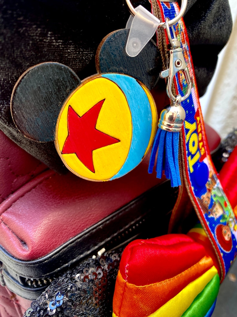 May include: A wooden keychain with a blue, yellow, and red design of a ball with a star. The keychain is attached to a brown lanyard with the words "Toy Story" printed on it. The lanyard has a blue tassel.