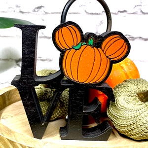 May include: Black wooden "LOVE" sign with a pumpkin Mickey Mouse head in the center. The sign is surrounded by small pumpkins and is sitting on a wooden surface.