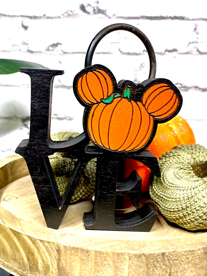 May include: Black wooden "LOVE" sign with a pumpkin Mickey Mouse head in the center. The sign is surrounded by small pumpkins and is sitting on a wooden surface.