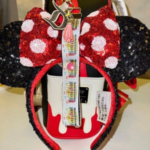 Pode incluir: Um la&ccedil;o de lantejoulas vermelho e branco com bolinhas com orelhas de lantejoulas pretas, preso a uma faixa de cabe&ccedil;a vermelha. Um cord&atilde;o com a palavra "Disneyland" est&aacute; preso ao la&ccedil;o. Uma pequena borla rosa e um pingente "D" tamb&eacute;m est&atilde;o anexados.