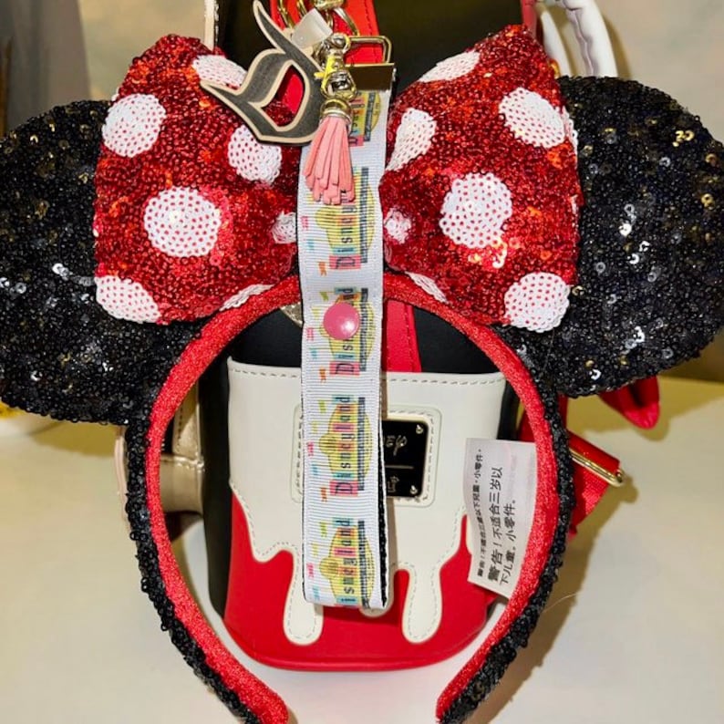 Pode incluir: Um la&ccedil;o de lantejoulas vermelho e branco com bolinhas com orelhas de lantejoulas pretas, preso a uma faixa de cabe&ccedil;a vermelha. Um cord&atilde;o com a palavra "Disneyland" est&aacute; preso ao la&ccedil;o. Uma pequena borla rosa e um pingente "D" tamb&eacute;m est&atilde;o anexados.