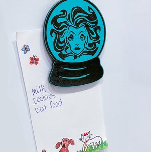 Op de afbeelding: Een turquoise blauwe sneeuwbol met een zwart silhouet van een vrouw met lang haar en een dramatische uitdrukking. De sneeuwbol is een houten ornament.