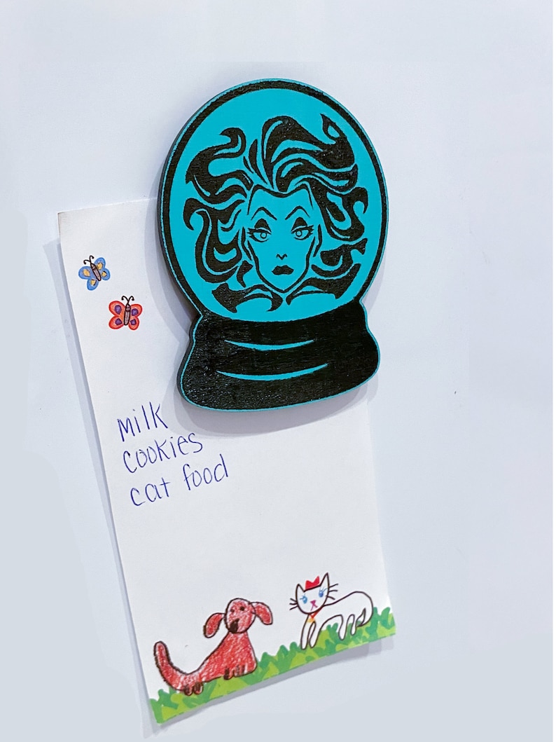 Op de afbeelding: Een turquoise blauwe sneeuwbol met een zwart silhouet van een vrouw met lang haar en een dramatische uitdrukking. De sneeuwbol is een houten ornament.