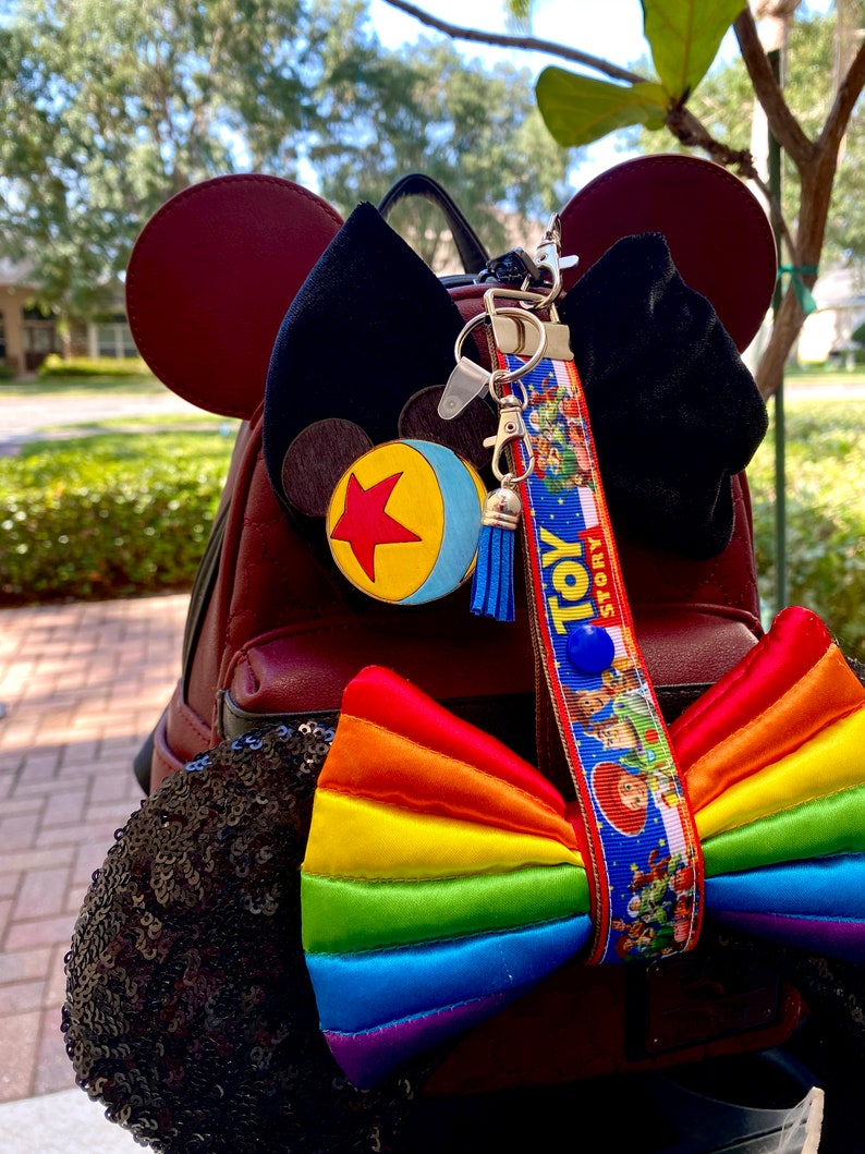 May include: A red faux leather backpack with black sequin ears and a rainbow bow. The backpack has a black velvet bow and a Toy Story keychain with a blue tassel.