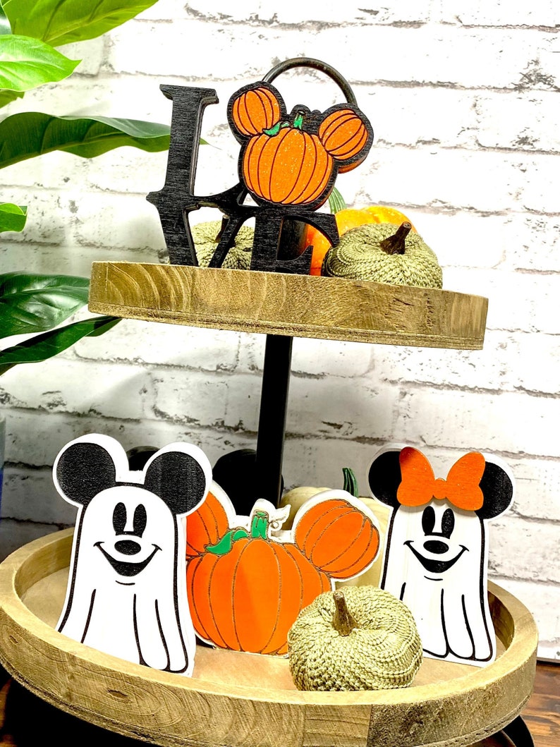 May include: A two-tier wooden tiered tray decorated with Halloween-themed decorations. The top tier features a black wooden "LOVE" sign with a pumpkin-shaped Mickey Mouse head. The bottom tier features two ghost-shaped Mickey Mouse and Minnie Mouse figures with pumpkins and gourds.