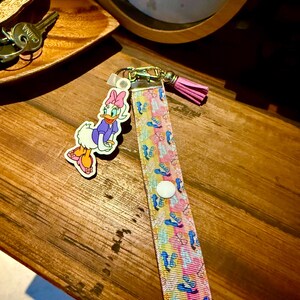 May include: A colorful lanyard with a Daisy Duck charm. The lanyard features a pattern of blue, pink, and yellow cartoon images. A pink tassel hangs from the gold-colored clasp. The Daisy Duck charm is a cartoon image of Daisy Duck wearing a purple top and pink bow.