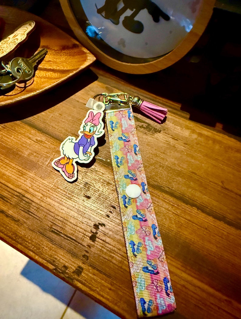 May include: A colorful lanyard with a Daisy Duck charm. The lanyard features a pattern of blue, pink, and yellow cartoon images. A pink tassel hangs from the gold-colored clasp. The Daisy Duck charm is a cartoon image of Daisy Duck wearing a purple top and pink bow.