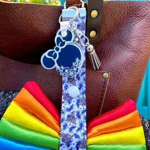 May include: A rainbow-colored satin bow with a white and blue floral patterned strap. The strap has a silver charm with a blue silhouette of Minnie Mouse's head with a bow. The strap is attached to a brown leather tote bag.