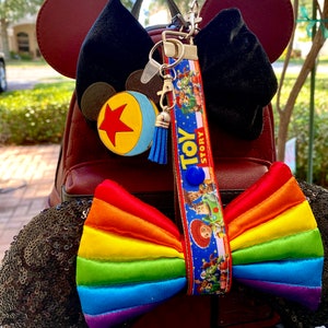 May include: A red faux leather backpack with black sequin Minnie Mouse ears. The ears have a rainbow bow and a Toy Story lanyard with a blue and yellow ball charm.