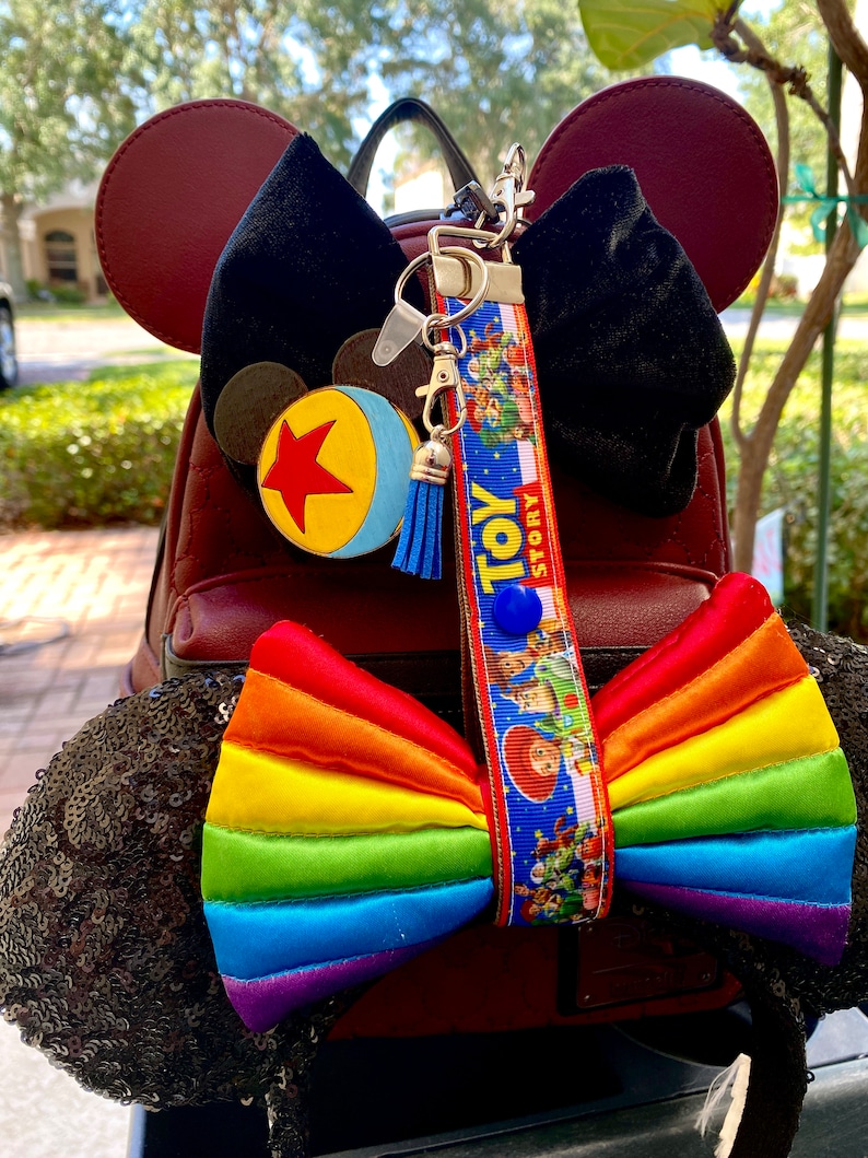 May include: A red faux leather backpack with black sequin Minnie Mouse ears. The ears have a rainbow bow and a Toy Story lanyard with a blue and yellow ball charm.