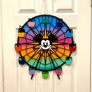 Puede incluir: Una noria de metal negro con un fondo degradado arco&iacute;ris y la cara de Mickey Mouse en el centro. La noria tiene 12 g&oacute;ndolas de colores.