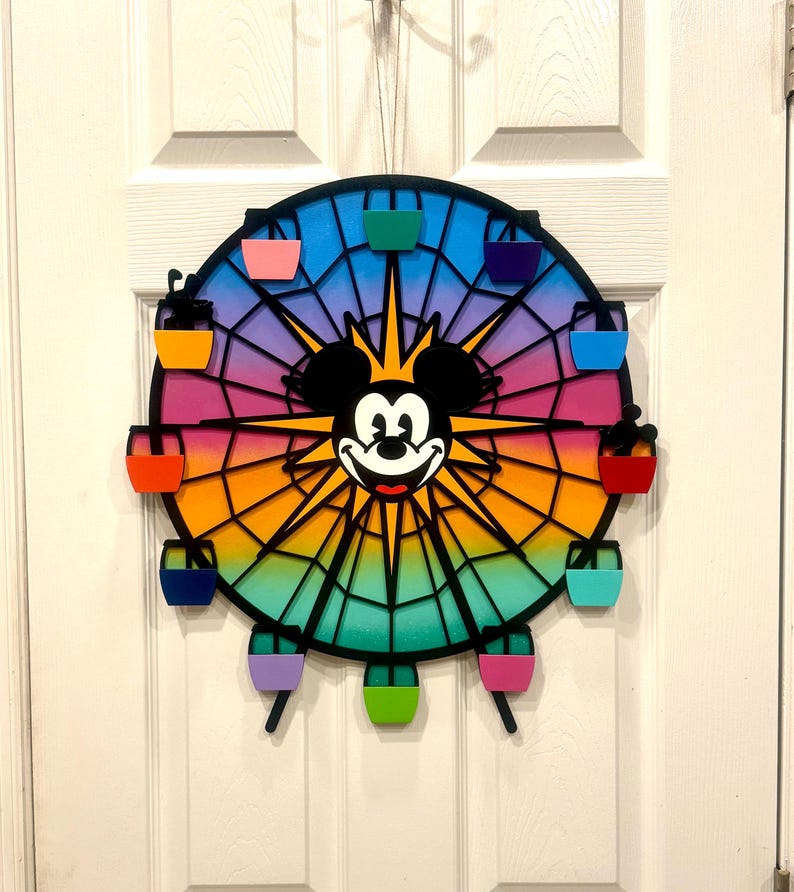 Puede incluir: Una noria de metal negro con un fondo degradado arco&iacute;ris y la cara de Mickey Mouse en el centro. La noria tiene 12 g&oacute;ndolas de colores.