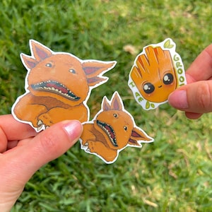 May include: Three cartoon character stickers. One is a brown, furry creature with a toothy grin. Another is a brown, furry creature with three eyes and a toothy grin. The third is a green, plant-like creature with a smiling face and the text "I am Groot".