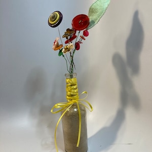Op de afbeelding: Een glazen flesvaas gevuld met zand en versierd met een geel lint. De vaas bevat een boeket van kleurrijke knoopbloemen met groene bladeren.