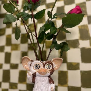 May include: A ceramic vase shaped like a Gremlin character, holding several stems of pink roses. The vase is white, brown, and tan with large eyes and a mischievous expression. The roses have dark green stems and leaves.