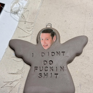 May include: A gray ceramic angel ornament with the text "I DIDN'T DO FUCKIN SHIT" carved into the body. A photo of a man's face is attached to the angel's head. The ornament has a metal loop for hanging.
