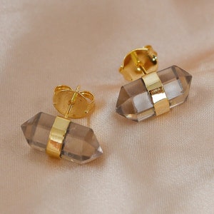 Pode incluir: Um par de brincos de ouro com pedras de cristal de quartzo transparente. As pedras têm a forma de um cristal de ponta dupla e têm uma banda dourada em torno do meio.