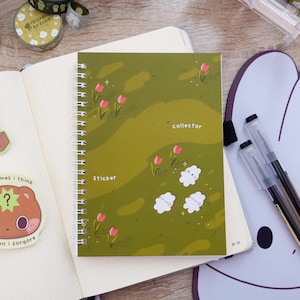 Op de afbeelding: Een spiraalgebonden notitieboek met een groene kaft met een veldontwerp met roze tulpen en witte wolkvormige vormen. De woorden "collector" en "sticker" staan op de kaft. Decoratieve washitape en stickers zijn ook zichtbaar.
