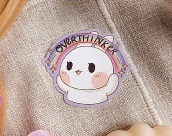 Puede incluir: Una pegatina blanca con un conejito de dibujos animados que lleva una diadema de arcoíris que dice "Overthinker". El conejito tiene ojos grandes y una nariz rosa.