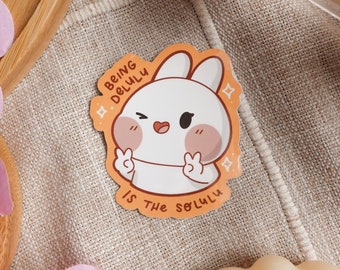 Puede incluir: Una pegatina con un conejo de dibujos animados con la nariz y las mejillas rosas. El conejo está guiñando un ojo y haciendo un signo de paz con ambas manos. La pegatina es naranja y tiene el texto "Being Delulu is the solulu".