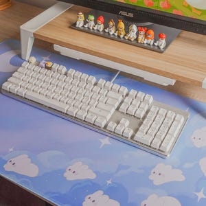 May include: A white mechanical keyboard with a silver frame sits on a blue mousepad with a cloud and moon design. A wooden monitor stand supports a computer monitor and small figurines.