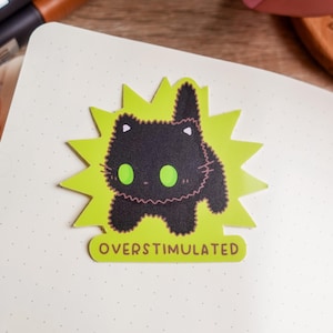Op de afbeelding: Een sticker met een zwarte kattenillustratie met groene ogen en een roze omtrek, tegen een limoengroene burst. De sticker heeft de tekst "OVERSTIMULATED" op een gele banner. De sticker ligt op een wit gestippeld oppervlak.